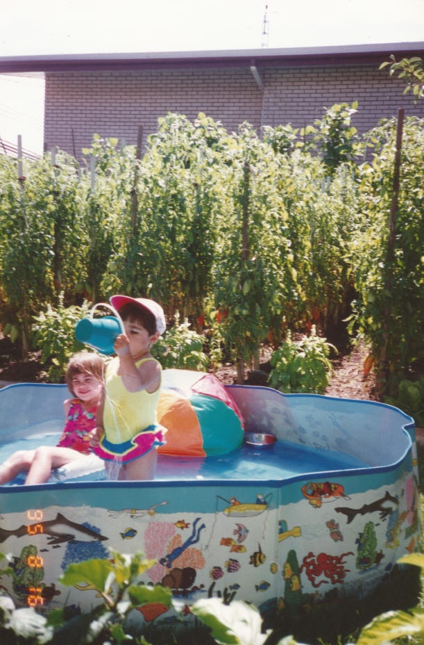 Two young girls in a kiddie pool. Behind them, are rows and rows of tomato plants. The photo is from 1995.