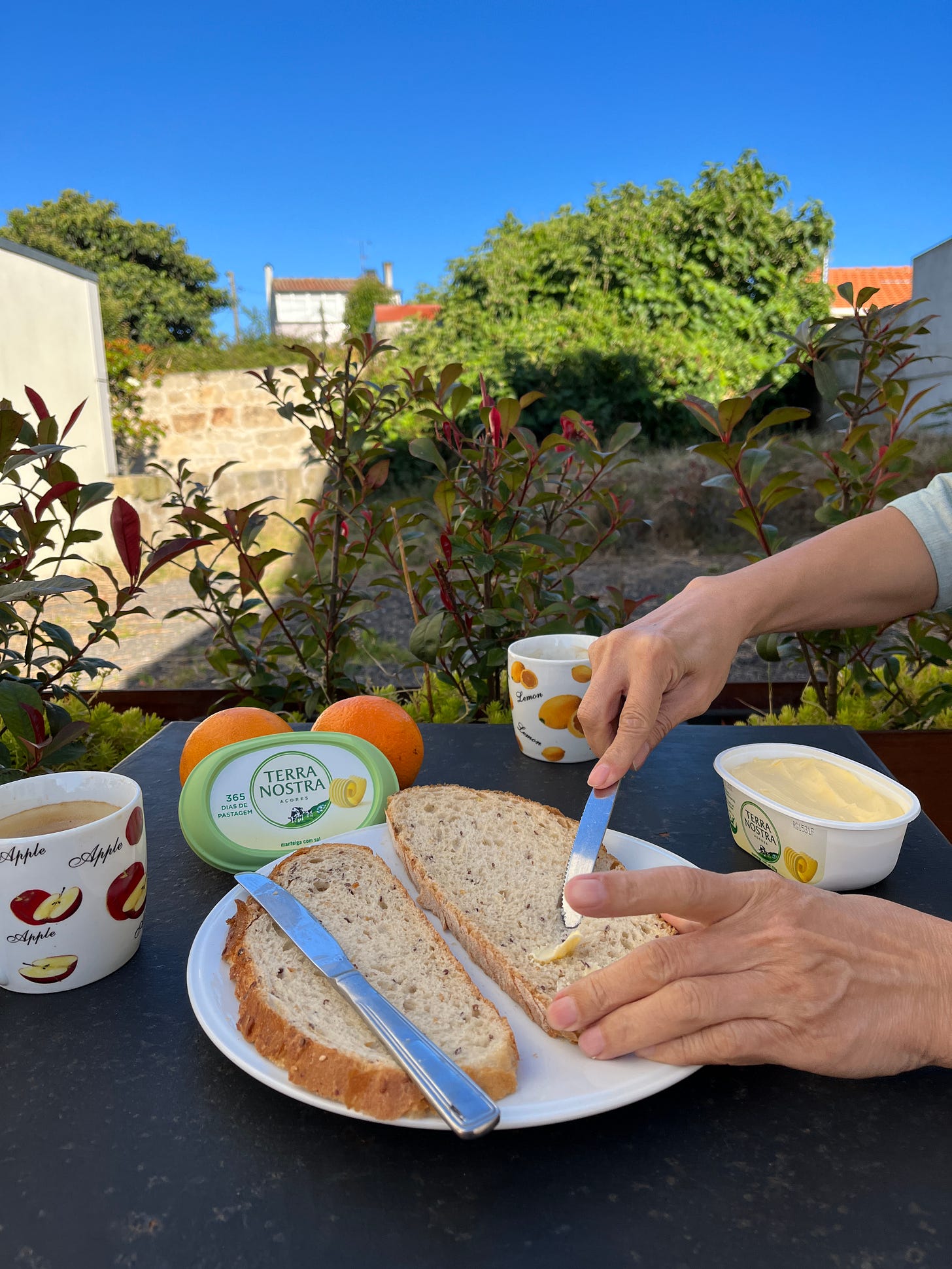 Image: photo of a breakfast scene, a small outdoor table with two plates of rustic bread, someone is buttering the bread, two mugs of coffee, two oranges, a tub of butter, the background are some trees and wild grasses; above, bright blue Mediterranean sky.