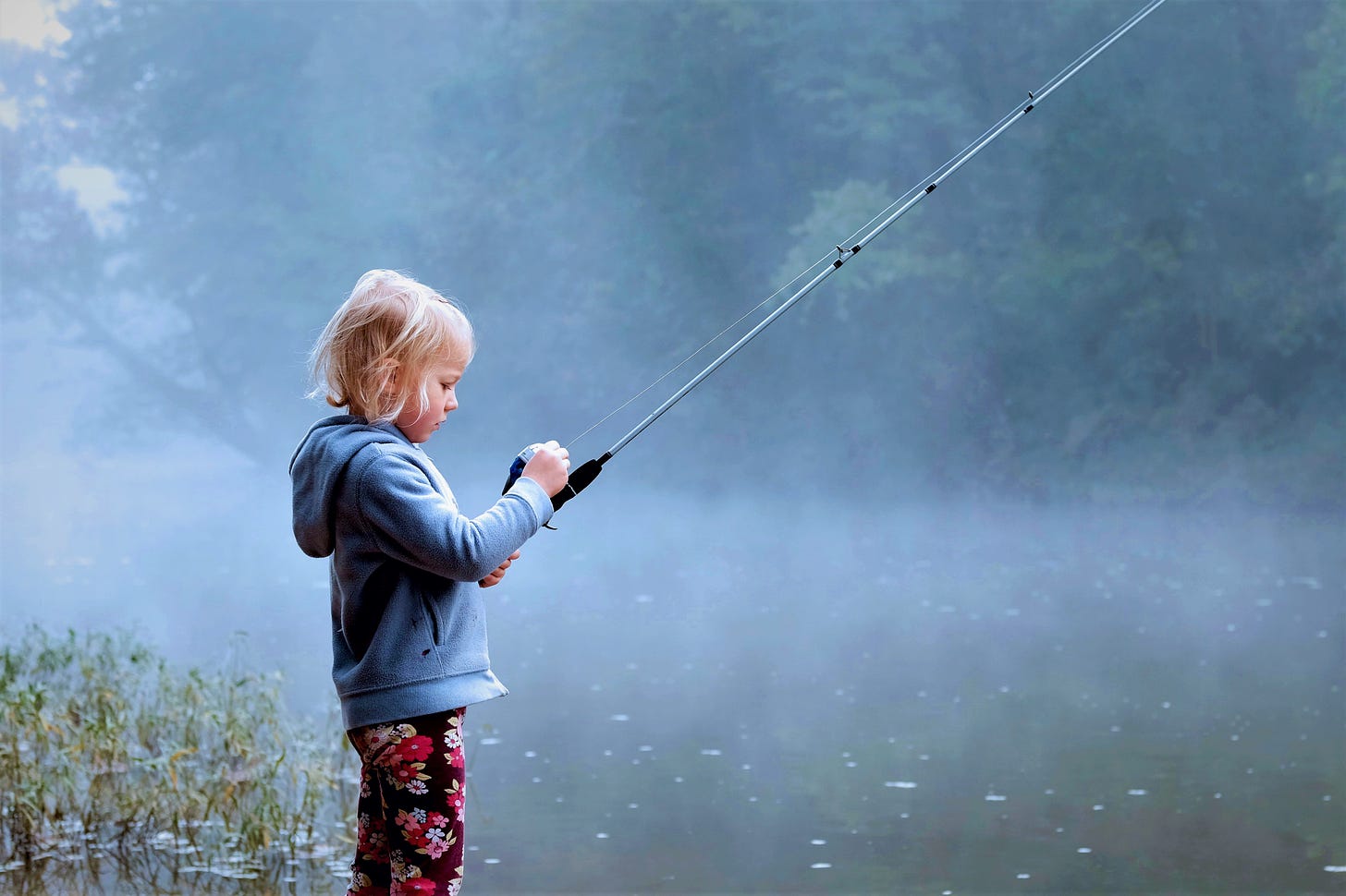 Young girl with blonde hair wearing blue sweatshirt fishing with rod and reel on quiet lake