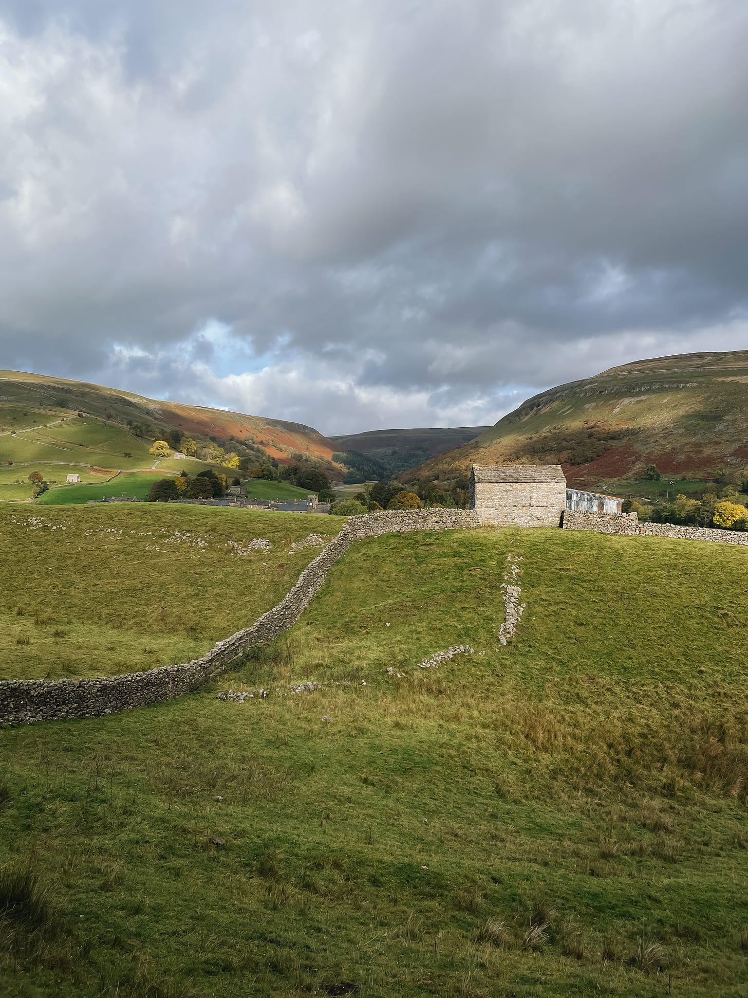 A landscape of hills and stone walls. A stone barn to the left of centre. Cloudy day.