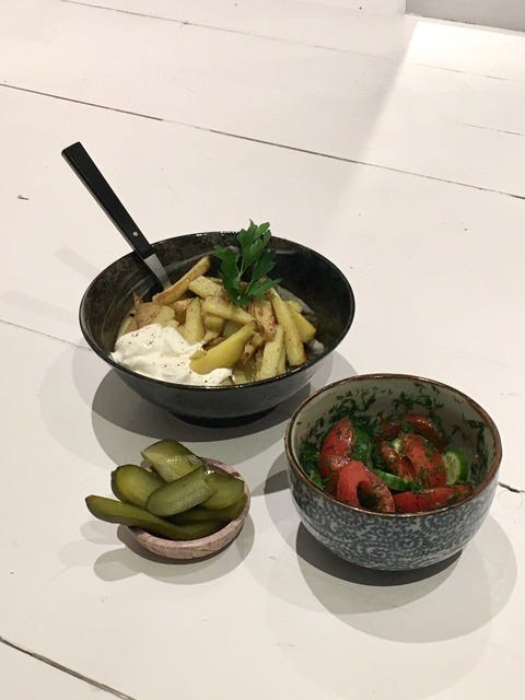 Three round dishes of different sizes; the largest with chopped fried potatoes, a dollop of sour cream, and a leafy garnish; the next, with rough chopped cucumbers and tomatoes flecked with dill; the smallest with thin green sliced pickles.  
