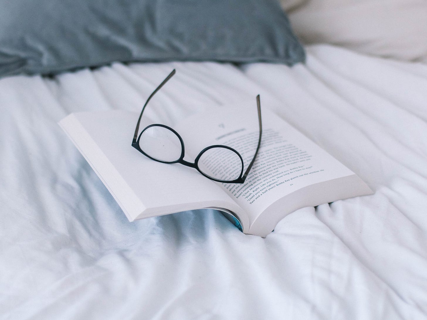 Photo of reading glasses laying on top of an open book that's laying on a bed.
