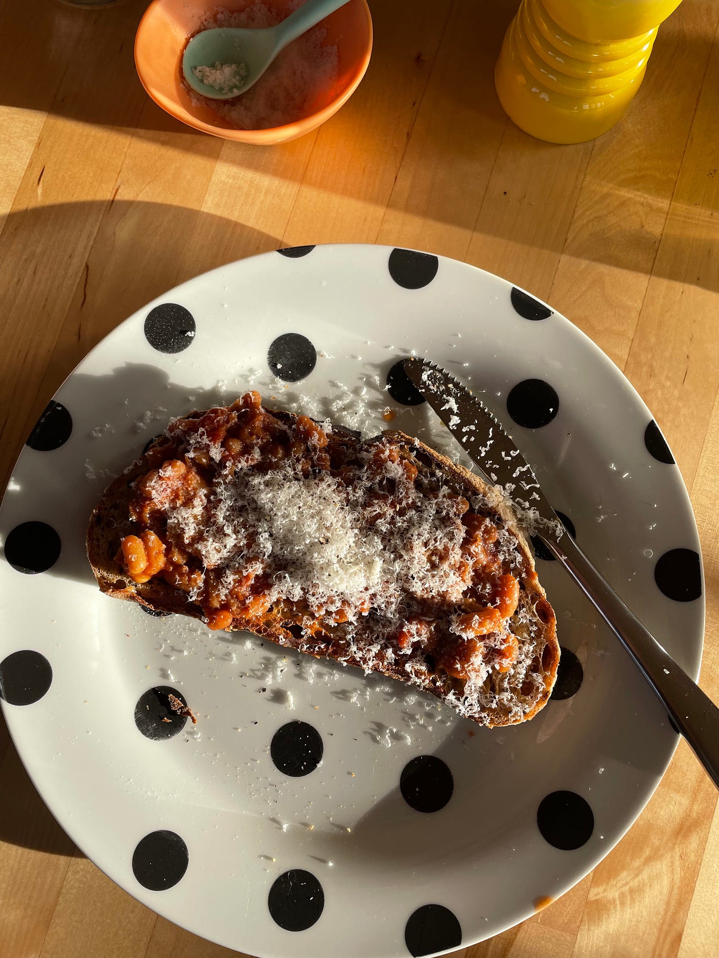 A piece of toast in the morning sun topped with baked beans and finely grated Parmesan cheese. Sea salt and a pepper grinder nearby. A warming any time meal.
