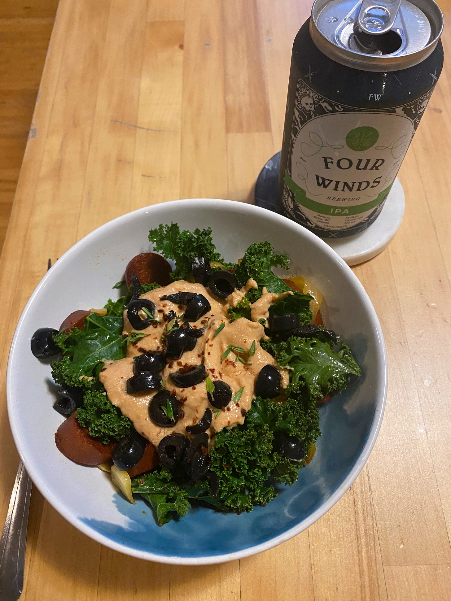 A white bowl full of rice, kale, browned slices of sausage, black olives, and an orange-ish cheezy sauce with sprinkles of thyme and chili flakes on top. On a coaster near the bowl is a blue can of Four Winds IPA.