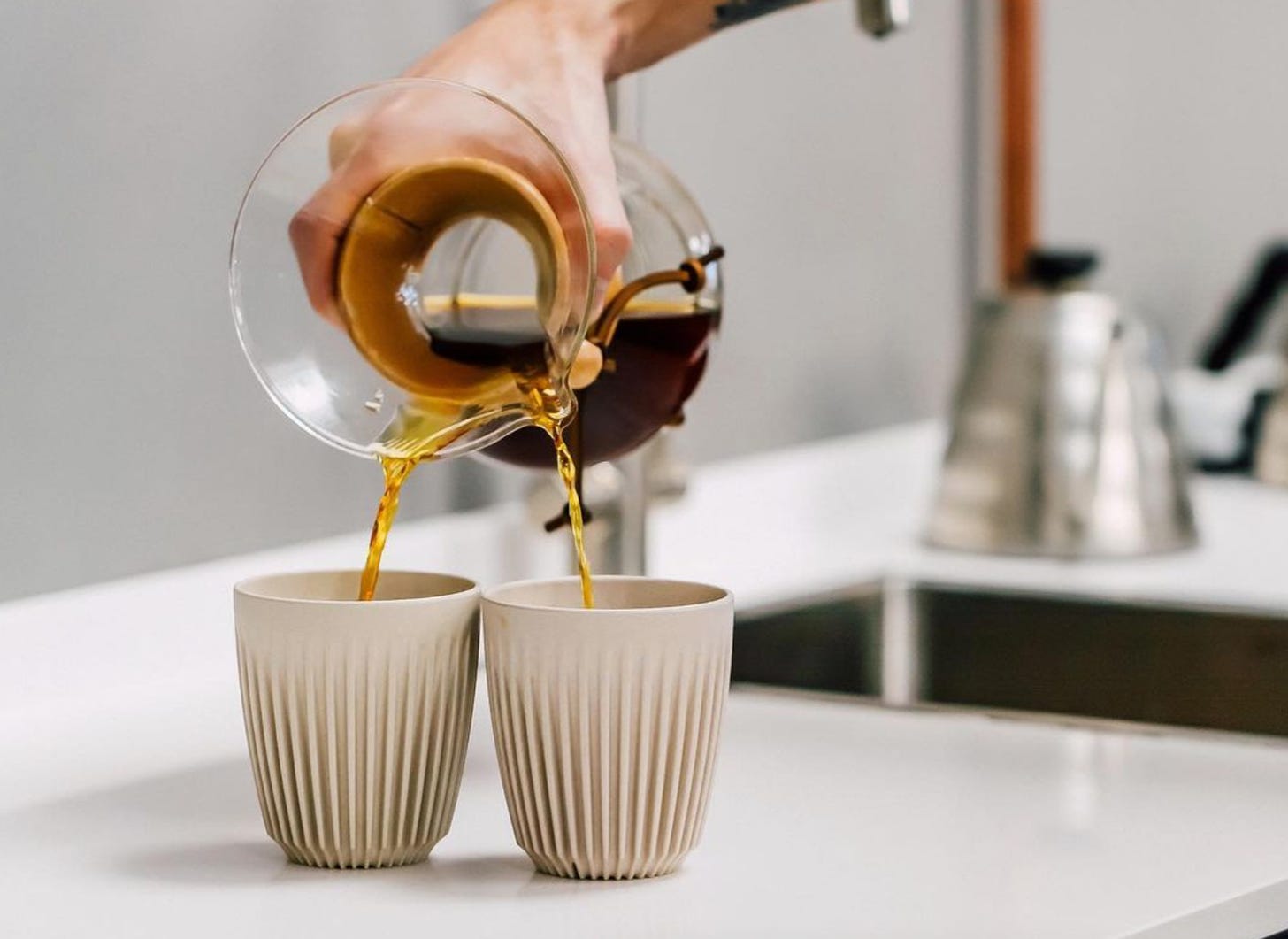 A close up on a hand pouring coffee from a full Chemex brewer pot into two beige cups simultaneously by angling the spout.