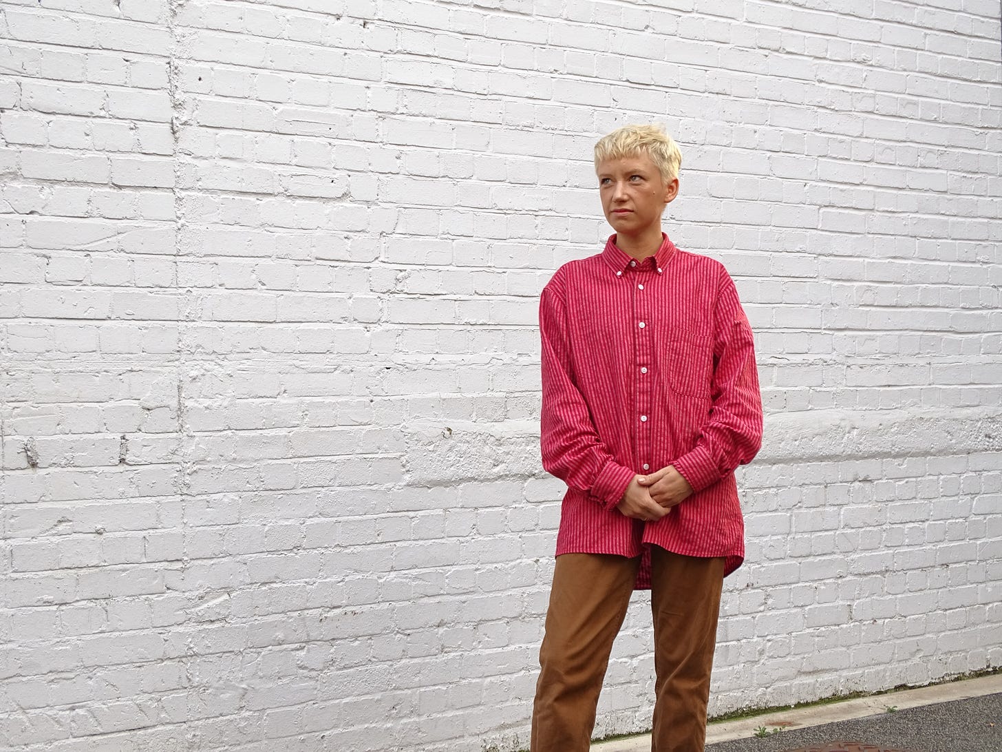 Photograph of author Alice Hattrick from the knee up, standing in front of a brick wall, wearing a striped button-down shirt with a cropped blonde haircut.