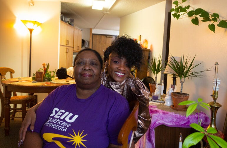 a Black woman with an updo hugs another Black woman sitting down from behind, in a living space near a dining table and floor lamp and next to a table with plants and figurines