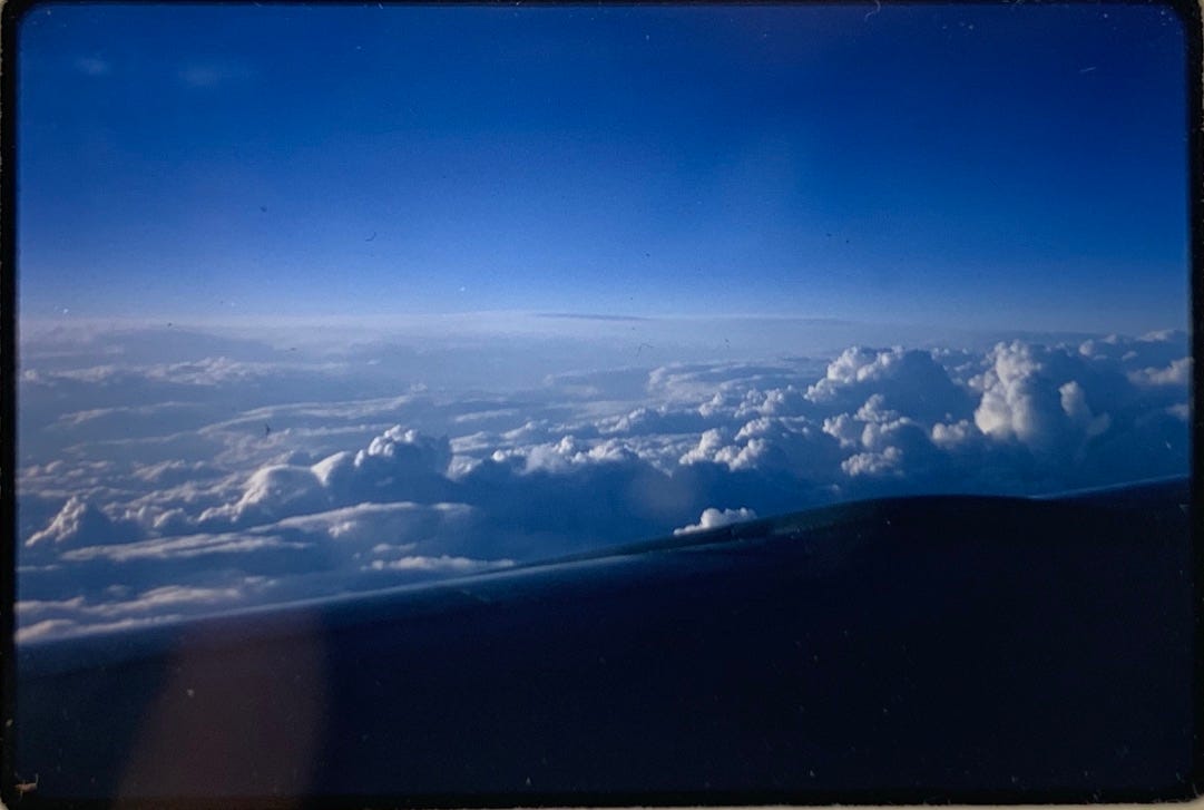 Photo of the view above clouds with an airplane wing visible in the lower half of the photo.