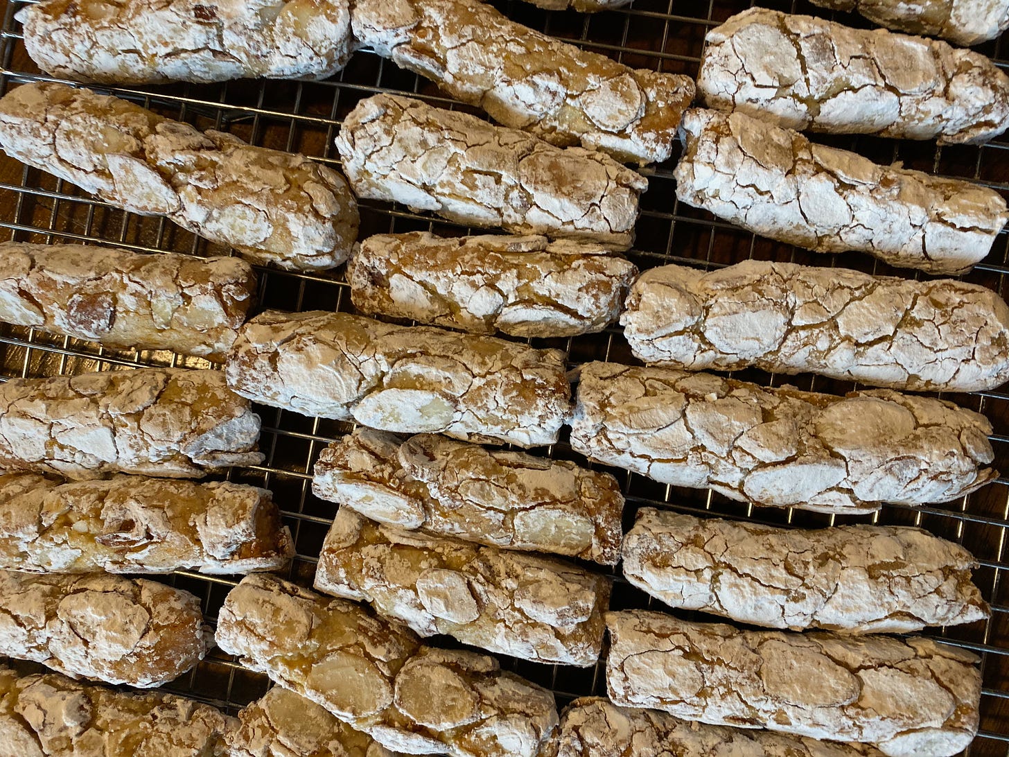Several rows of small cylindrical, log-shaped cookies sit on a metal cooling rack. They have flaked almonds pressed into them and are coated in powdered sugar.