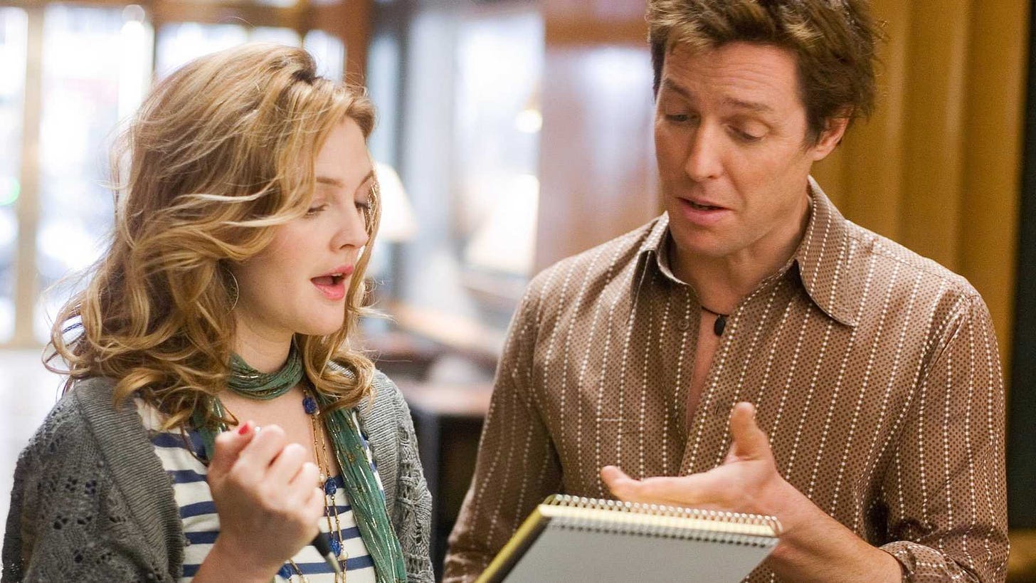A white woman, played by Drew Barrymore, holds a pen and shows a notepad to a white man, played by Hugh Grant