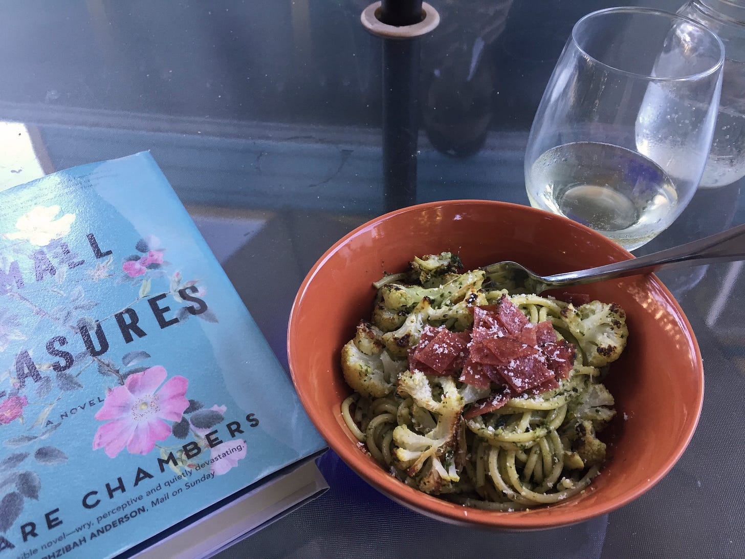 An orange bowl of pesto and cauliflower spaghetti with parmesan and pieces of salami on top sits between a blue hardcover book and a stemless glass of white wine. The book's title is "Small Pleasures".