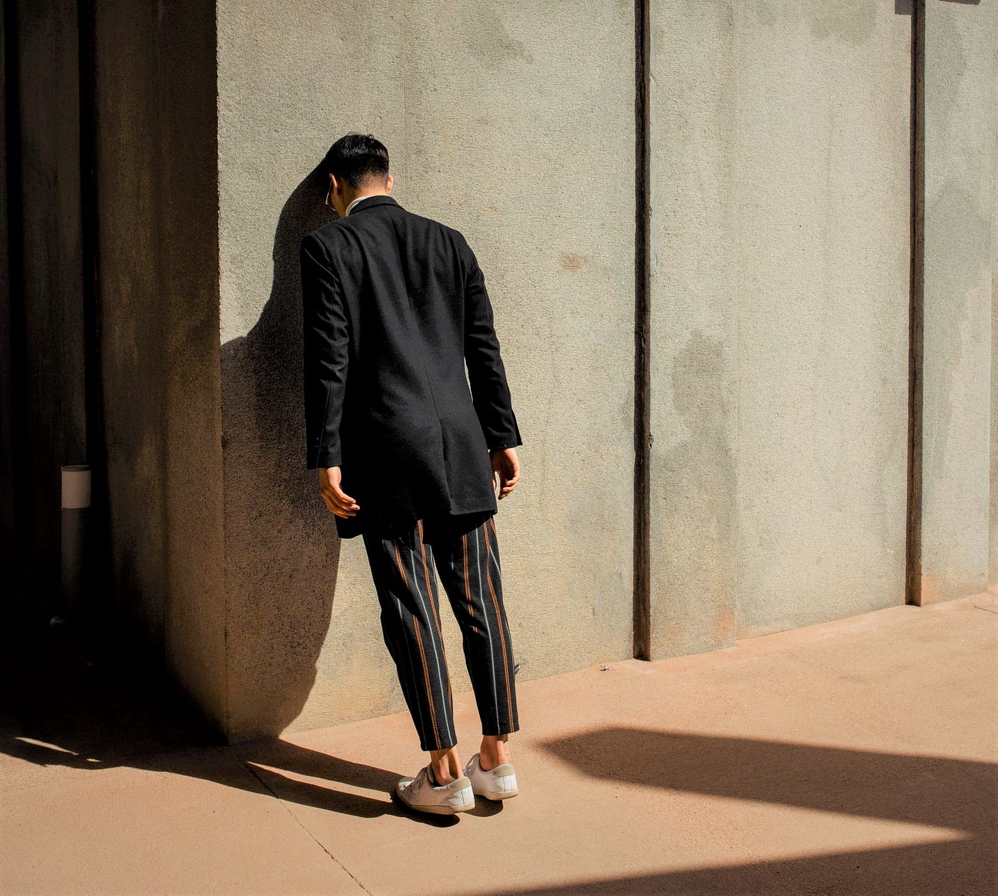 man wearing long sleeve dark top and striped pants leaning head against concrete wall