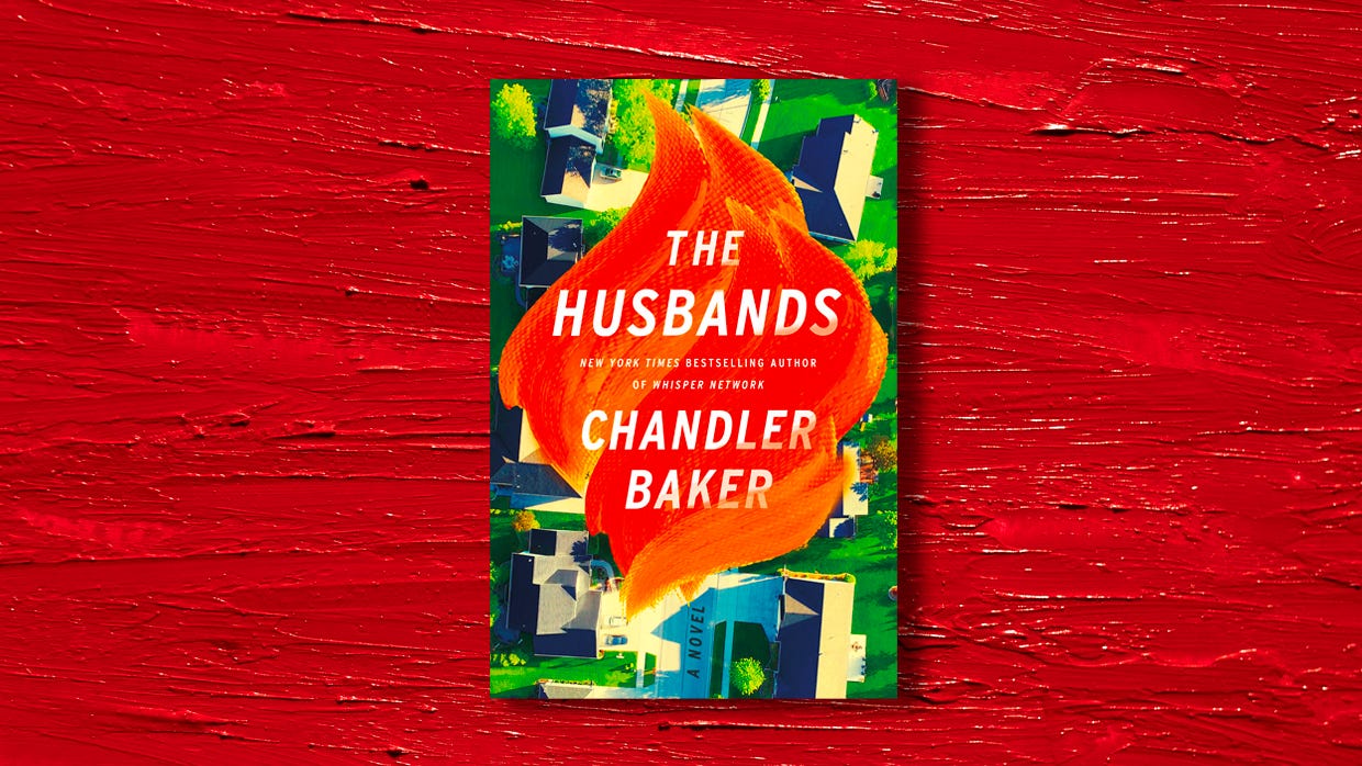 A copy of the book The Husbands by Chandler Baker lying on a red wooden surface. The cover shows a suburban neighborhood over which big orange flames float.