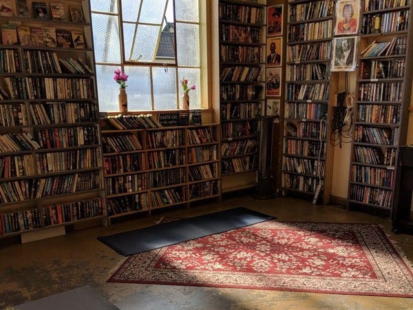 A room lined with bookshelves. A red rug is on the ground, and on top of that, a black yoga mat. Sunlight pours through a large window on the left wall. 