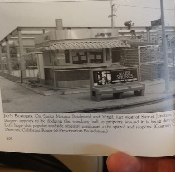 A photo of a picture + caption in a book, being held by someone whose hand is visible holding the page open. The image in the book is black and white and shows a very old version of Jay’s where the shack looks all metal with an awning and some windows. There’s a bus bench in front of the shack and a parking lot behind it. 