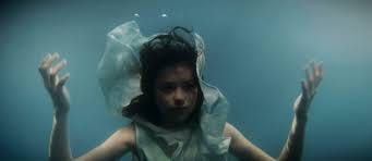 Cena do filme "A Chorona". Uma menina de cabelos escuros, embaixo d'água, ergue os braços. A gola de seu vestido está subindo com o movimento das águas.