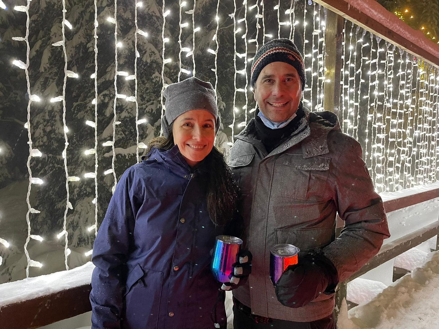 Two people in big puffy coats on a lighted bridge path in the snow