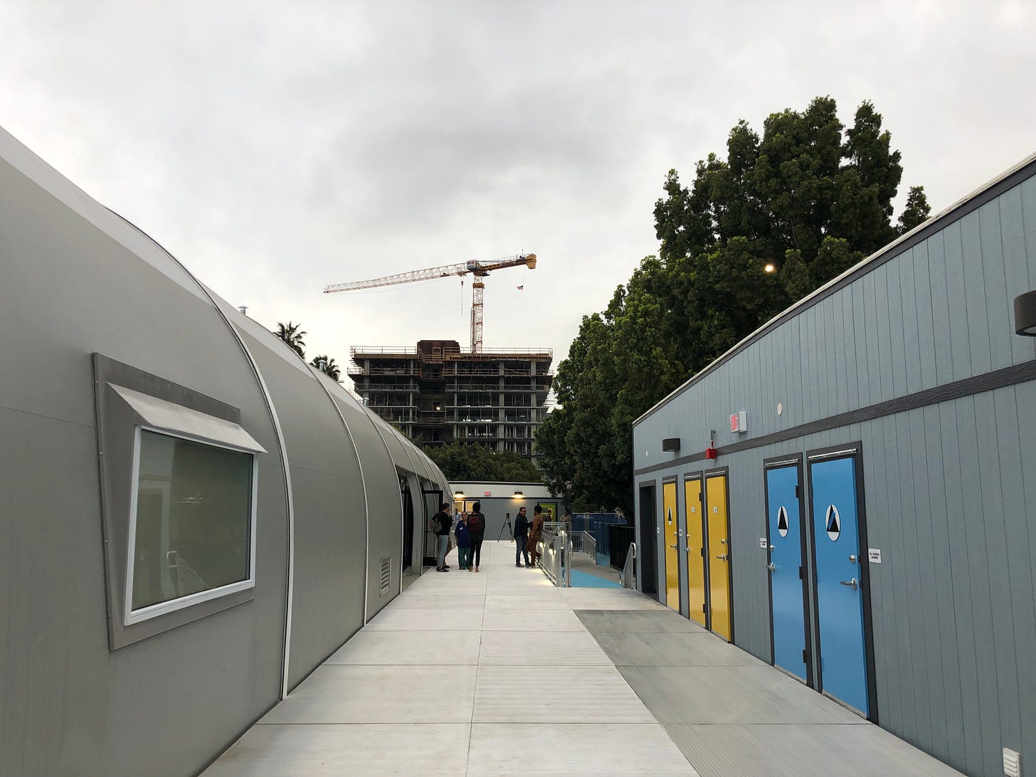 The exterior of the Schrader shelter, showing a walkway between the large tent and bathrooms in a portable building. In the background, a construction crane builds a high rise apartment building.
