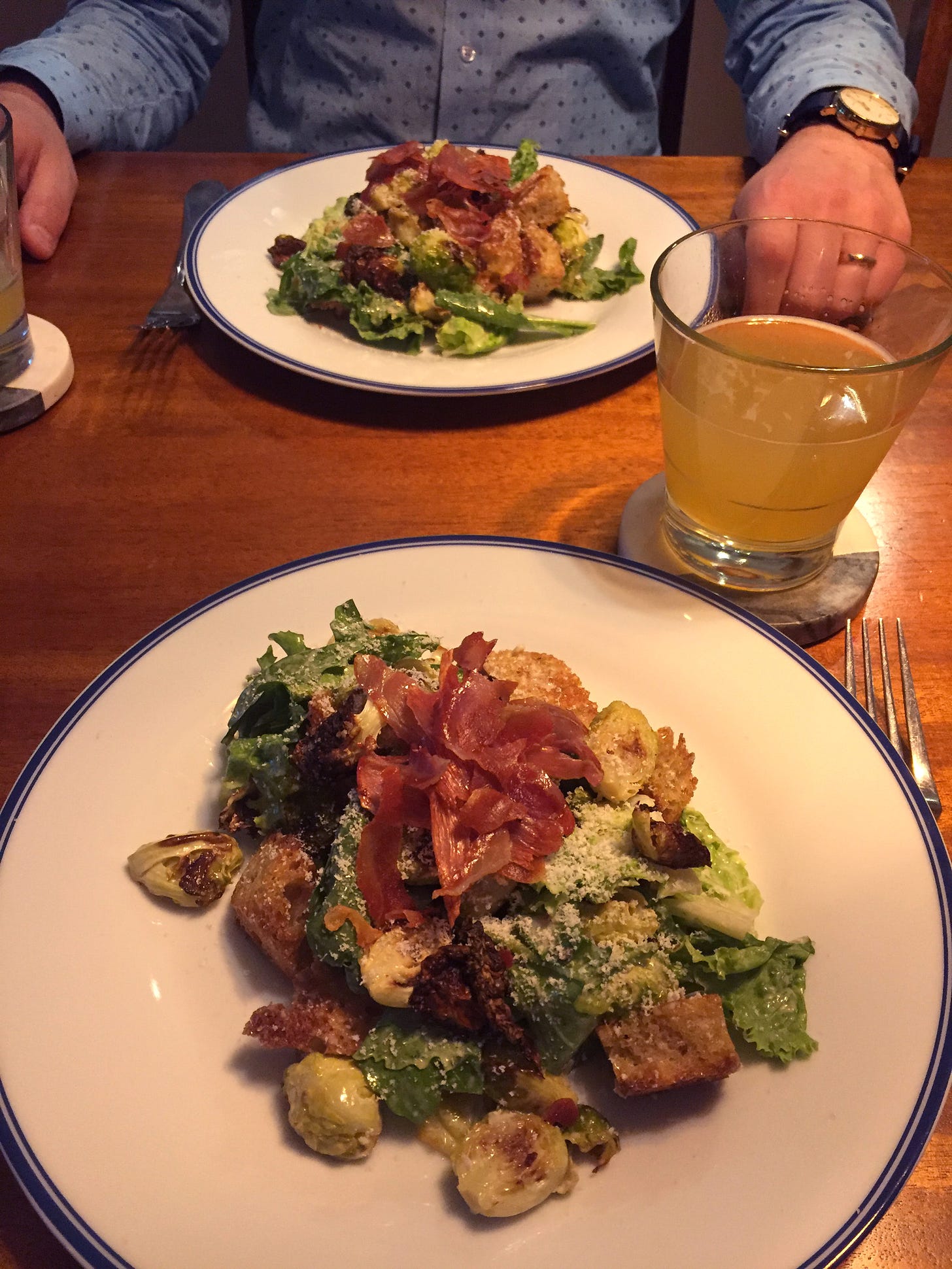 Two plates across from each other at the table, each with a serving of Caesar salad topped with pieces of prosciutto and finely grated parmesan. Large croutons and charred, roasted brussels sprouts are visible throughout the salad.