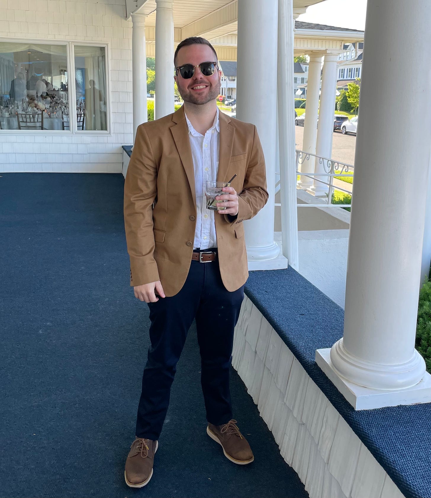 A thirty-something white male in sunglasses and a brown suit jacket stands outside of a wedding holding what appears to be a vodka tonic. A thirty-something white male in sunglasses and a brown suit jacket stands outside of a wedding holding what appears to be a vodka tonic.
