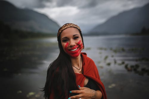 [ID: woman stands in front of dark, blurred background of grey mountains, clouds, and a wide river. She is smiling, has long, dark, brown hair and light brown skin, wearing a red cape and headband, fingernails painted teal with a tattoo on the back of her hand, and a red handprint painted across her face.]