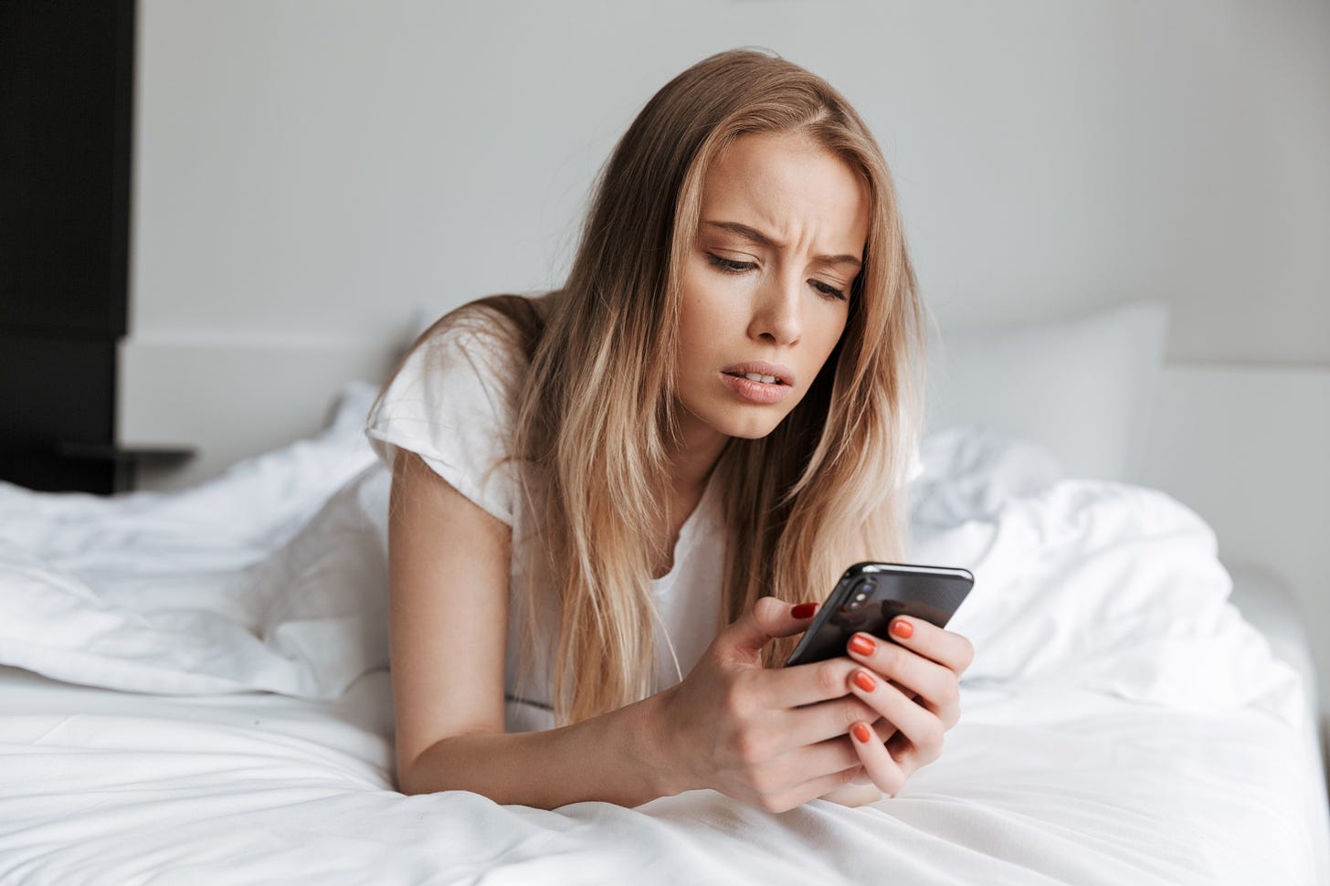 An upset woman stares down at her phone while she lies in bed.