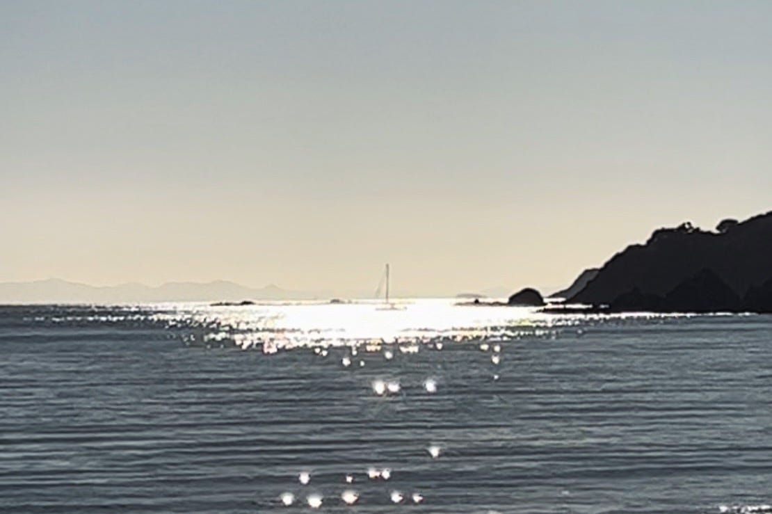 A poor-quality, blurry photo of an ocean scape with a central concentration of reflected sunbursts on the water and a yacht just visible in the muddle of the reflections. To the left are small layered peninsulas and in the foreground the ripples of the water are visible