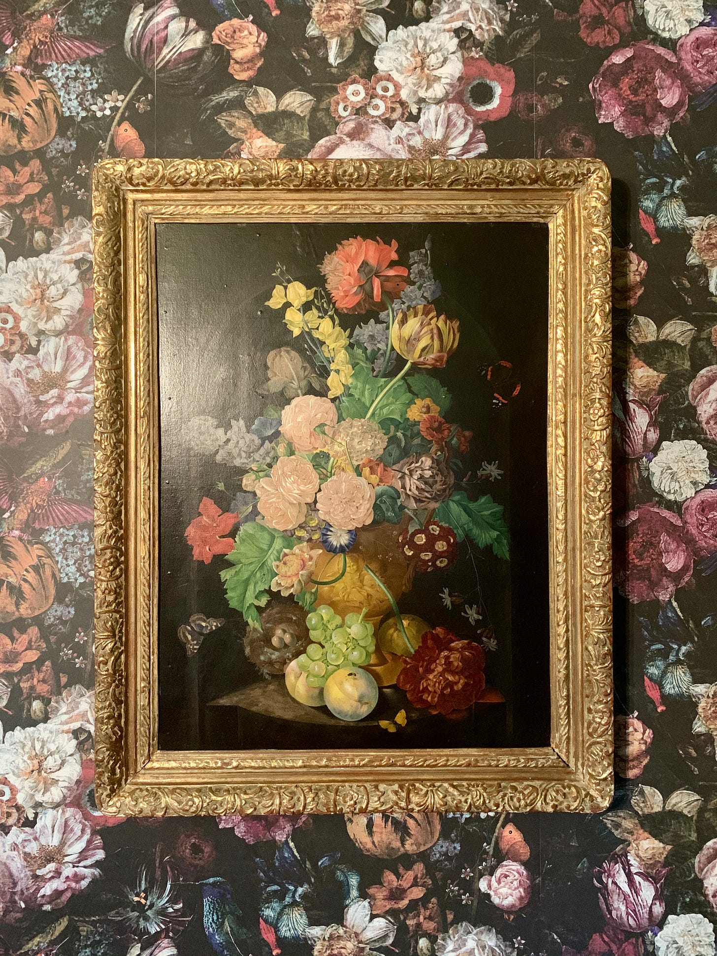 A floral still life in a gold frame hung on floral wallpaper