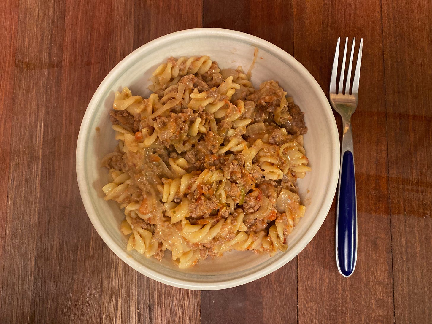 A white ceramic bowl filled with fusilli pasta tossed with a lamb tomato sauce. A fork sits on the wooden counter next to the bowl.
