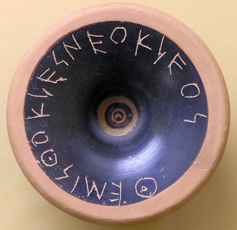 A round, orange and black piece of clay, on which Themistocles is scratched in Ancient Greek.