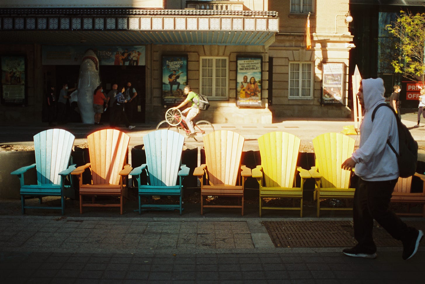 On a sidewalk, a row of multi-color Muskoka chairs, with a man in a grey hoody in the foreground; in the background a man is riding a bike and holding a third wheel, and behind him a crew is moving a ceramic shark statue to the front of a theater promoting a musical about Jaws, the movie.