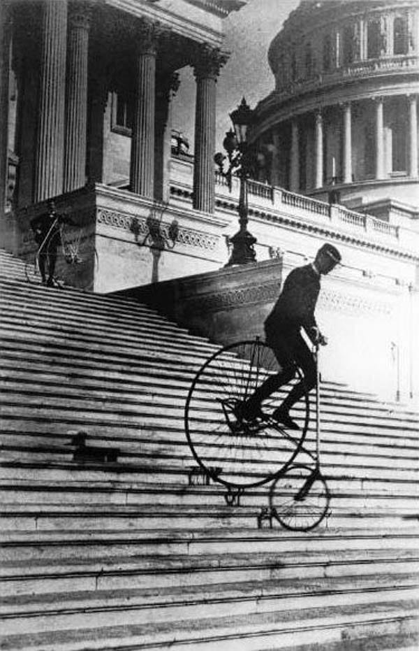 Black and white photo of Will Robinson, wearing dark slacks and jacket and a cap, riding a bicycle down the steps of the US Capitol. Robinson is on a bike with a small (around 20" diameter) front wheel and a large (around 50" diameter) rear wheel. Robinson sits on a seat located above the giant rear rear with his feet on pedals situated near the axle of the rear wheel, and gripping a narrow handlebar that is connected by a long rod to the front wheel. The bike resembles a penny farthing in reverse.   Behind Robinson, up the steps, stands another man besides his own bicycle, watching Robinson descend. Beyond him are a colonnade of Corinthian columns and beyond that is the dome of the Capitol building.
