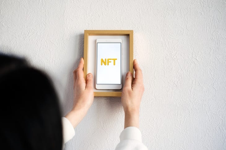 Image of a woman placing a framed phone with "NFT" on the screen against a white wall.