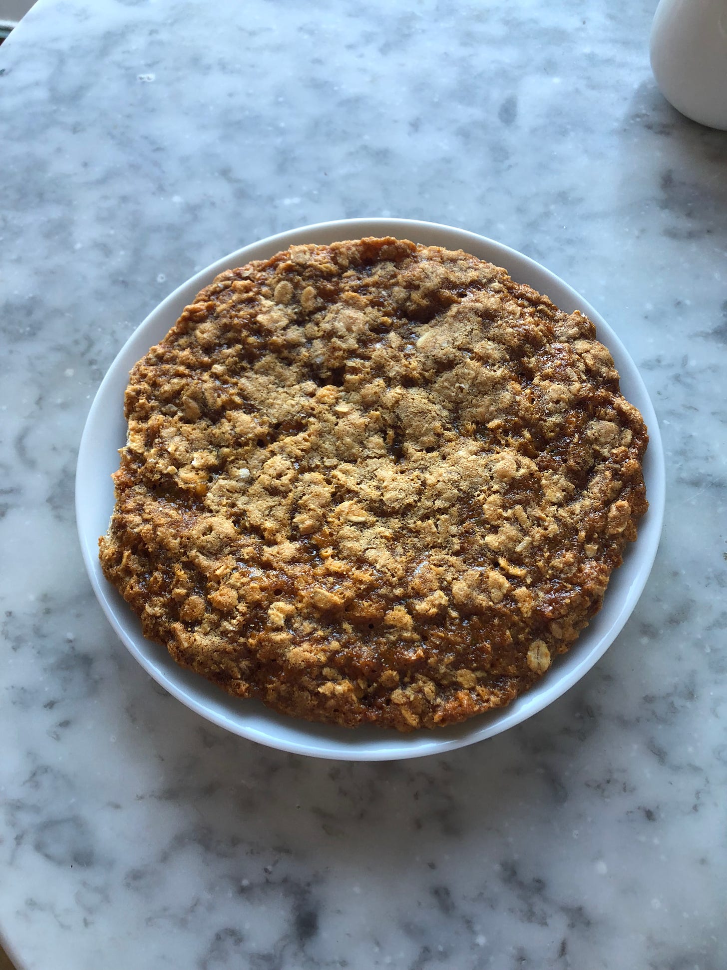 A wobbly brown oaty Anzac cookie sits on a saucer the perfect size for it, on a mottled marble table. 