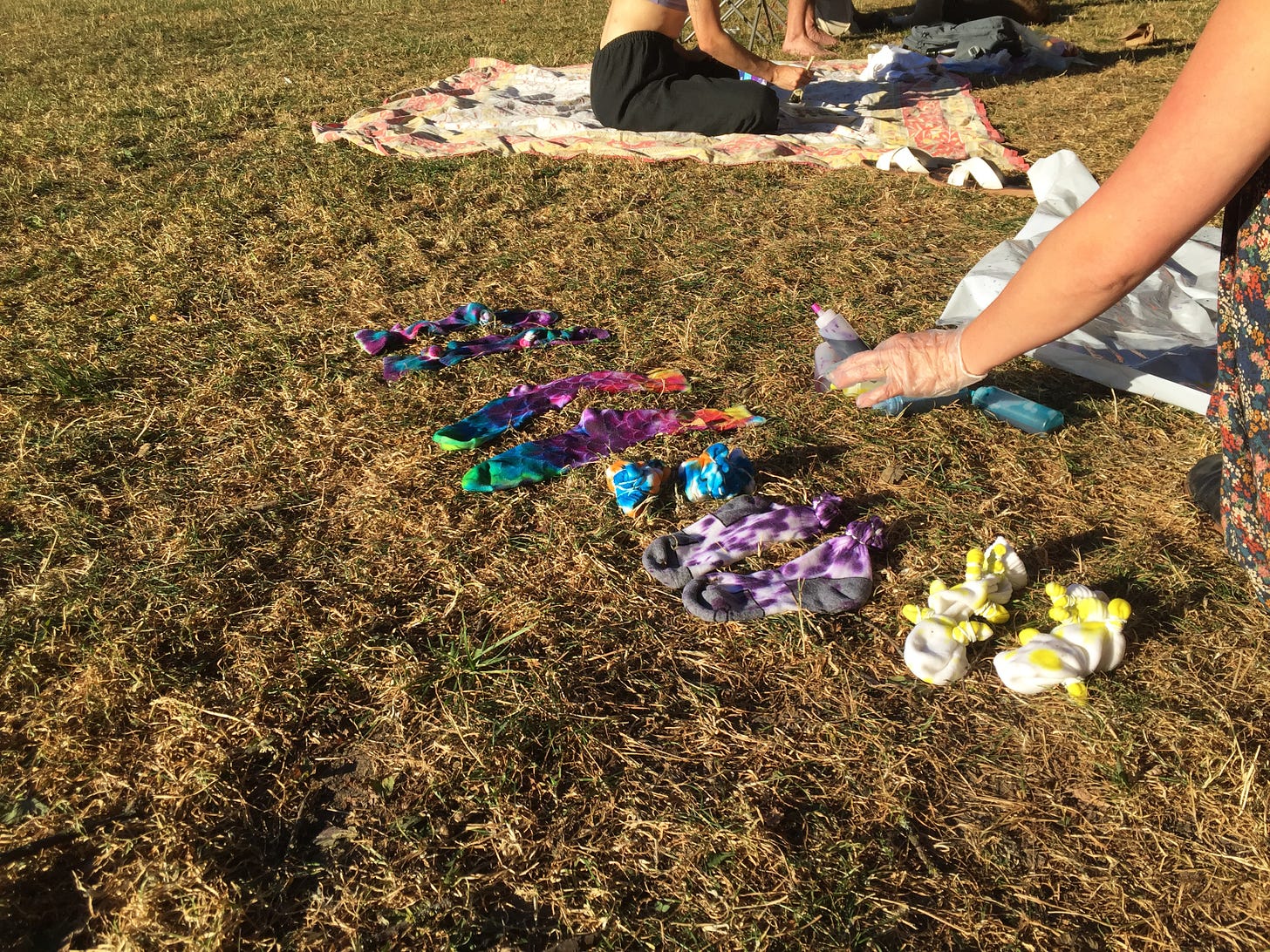several pairs of tie-dyed socks lay on grass that looks dry and yellow. A gloved hand holding a bottle of dye is in the right side of the photo. In the background, people's legs are visible sitting on a picnic blanket.