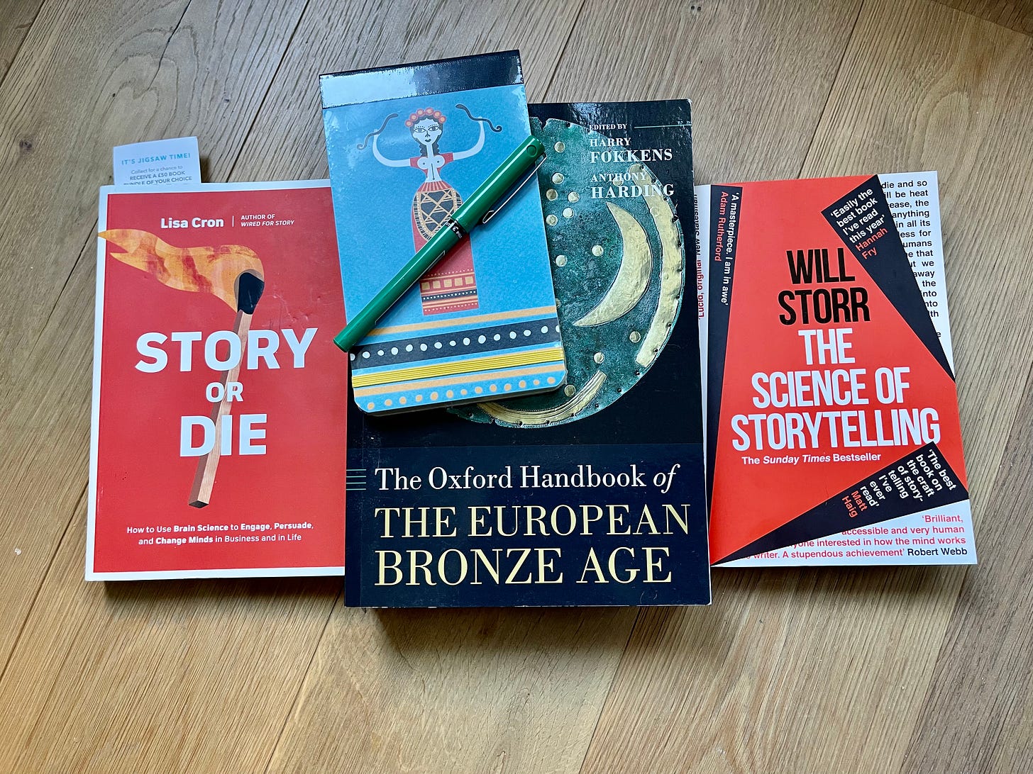 A hardwood floor with three books: Story or Die, The Oxford handbook of the European Bronze Age and The Science of Storytelling. On top of the books is a little notebook and a green pen