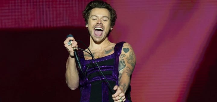 A man in purple velvet overalls with a microphone and mic stand, mouth open, eyes closed