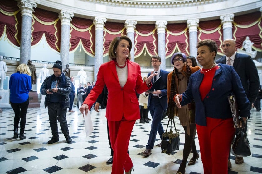 Nancy Pelosi, in a red pant suit, strides through the National Statuary Hall in the US Capitol. She is walking alongside a black woman. They are being trailed by aids. In the background, the columns of the round hall can be seen with burgundy fabric draped between them.