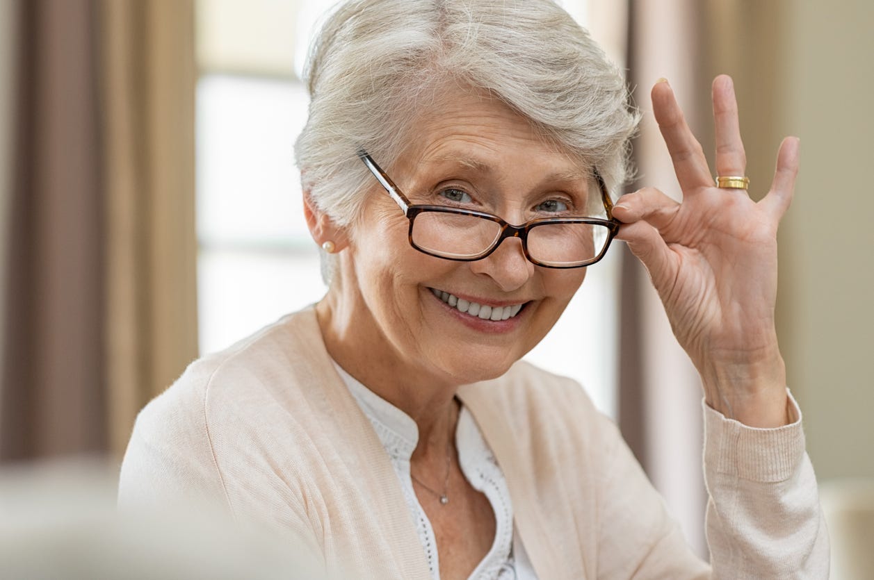 Smiling older woman with gray hair as she tips her glasses below her bright eyes with her left hand.