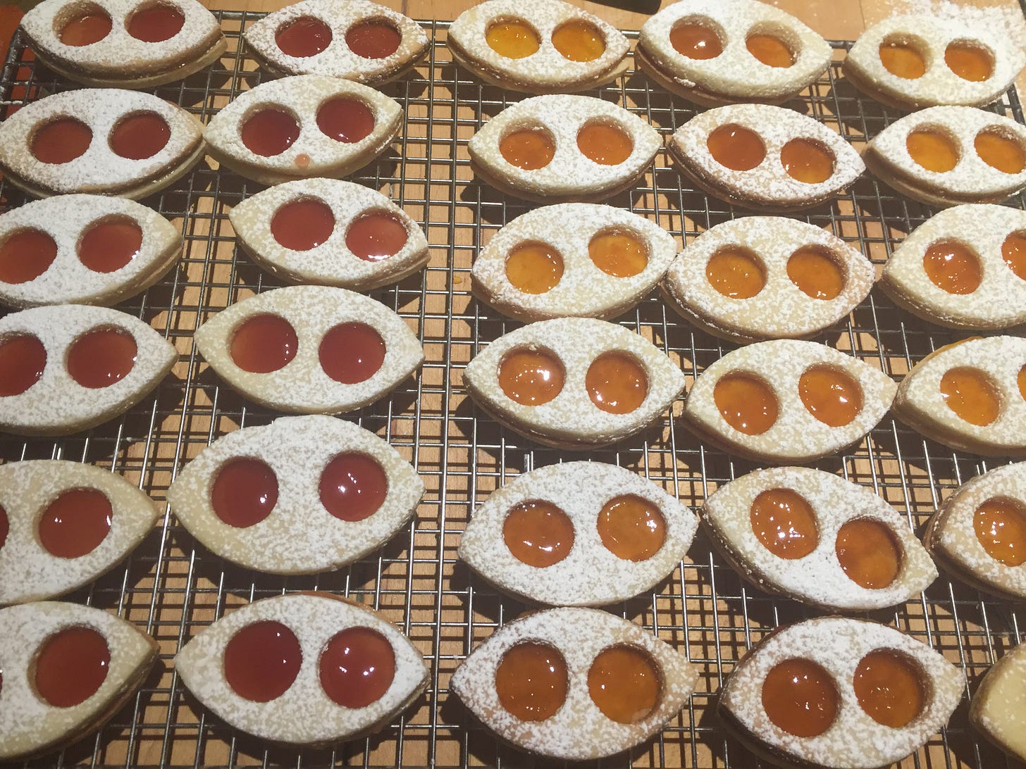 Many rows of teardrop shaped sandwich cookies. Each cookie has two round holes cut out of it, through which you can see the jam filling. Half are filled with a deep red raspberry jam; the other half with golden apricot jam.