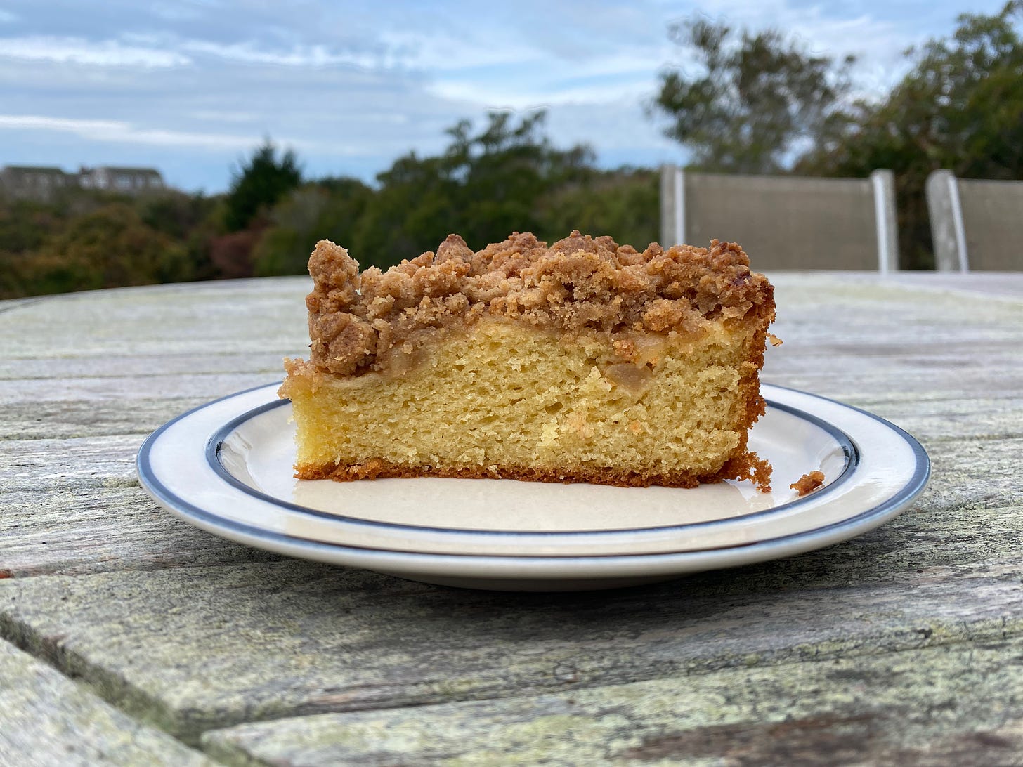 A tall slice of cake, topped with pear slivers and a brown sugar crumble, sitting on a plate on an outdoor table.