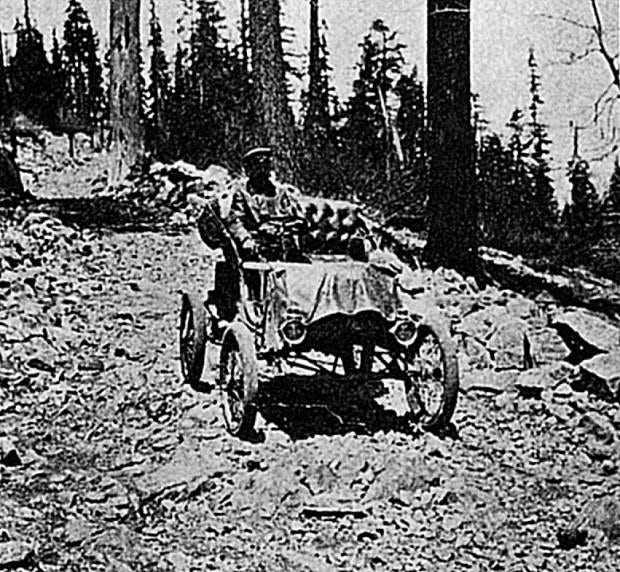 Grainy black and white photo of Alexander Winton driving one of his own cars over Donner Pass. The car is heading down a small incline with tall evergreens in the background. The "road" is more of a rocky path, with fist-sized rocks on the path and larger boulders to the sides. The car is very simple, 4 narrow spoked wheels attached to a simple chassis with two round headlights and fabric draped over the hood area. Winton, wearing a cap and coat sits high on an upholstered seat, on the right hand side the car where the steering wheel is located. 