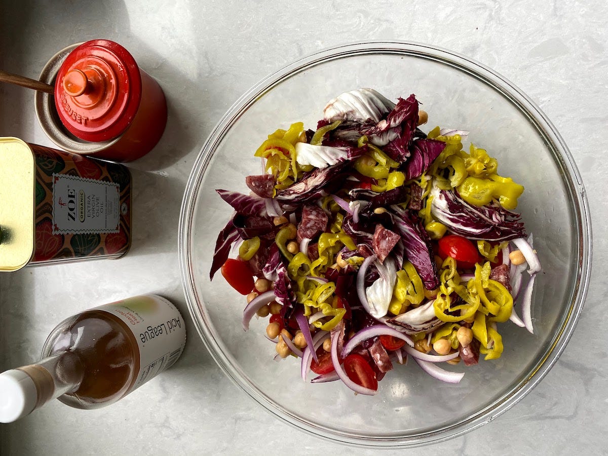 An overhead view of a glass bowl containing radicchio, pepperoncini, tomatoes, chickpeas, red onion, tomatoes and salami. Olive oil and a bottle of vinegar sit to the left of the bowl.  