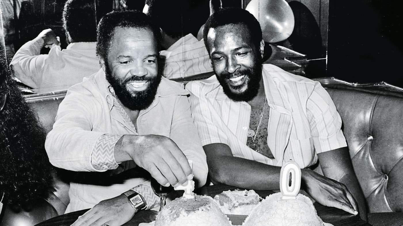This image shows Motown founder Berry Gordy and Marvin Gaye sitting in a restaurant booth while celebrating Gaye's 40th birthday. Gordy sits on the left and grasps a candle shaped like the number "4" sitting atop a birthday cake. Gaye, sitting on the right, smiles as Gordy does so. This image shows Motown founder Berry Gordy and Marvin Gaye sitting in a restaurant booth while celebrating Gaye's 40th birthday. Gordy sits on the left and grasps a candle shaped like the number "4" sitting atop a birthday cake. Gaye, sitting on the right, smiles as Gordy does so.
