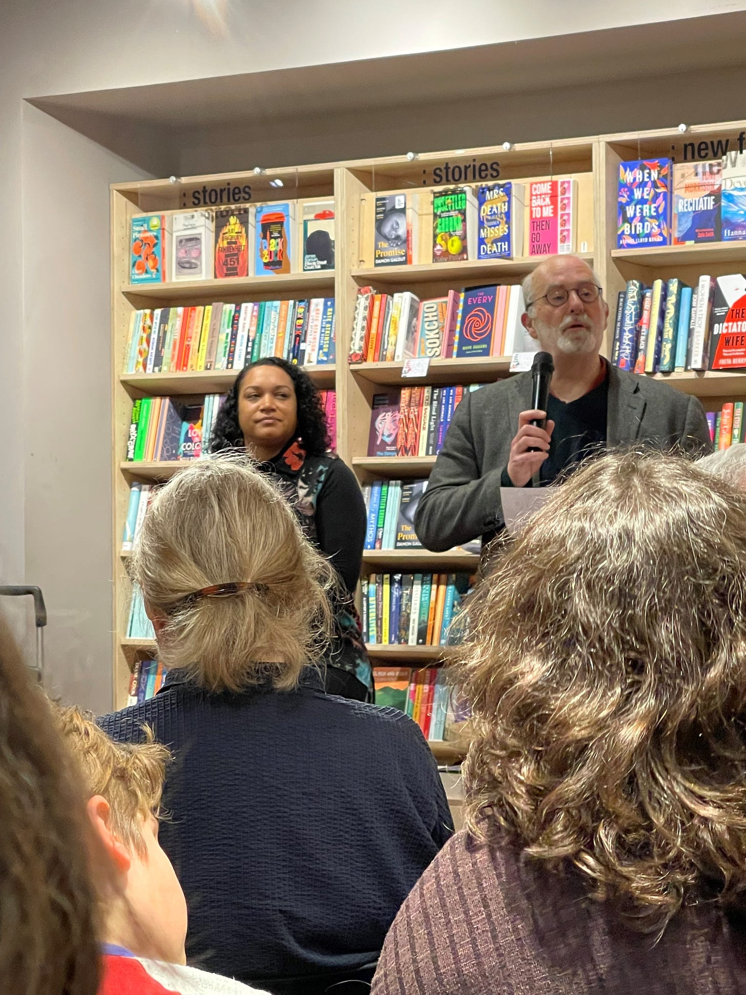 Inside Bookhaus, Clive Stevens and Cleo Lake standing in front of a set of book shelves, an audience about 50, sitting in front. Clive speaking.