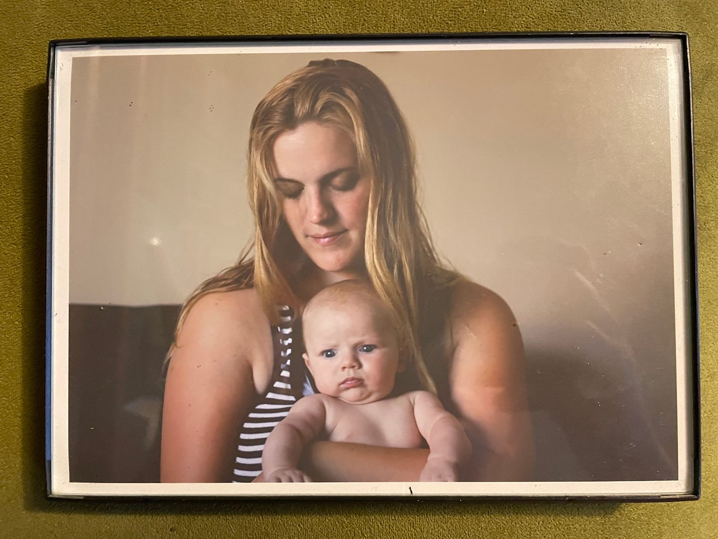 DL is looking down while holding a grumpy 5 month old baby who is very cute. DL's eyes are downcast but she has a very slight smile on her face. Her hair is slightly wet and she is wearing a black and white striped tank top