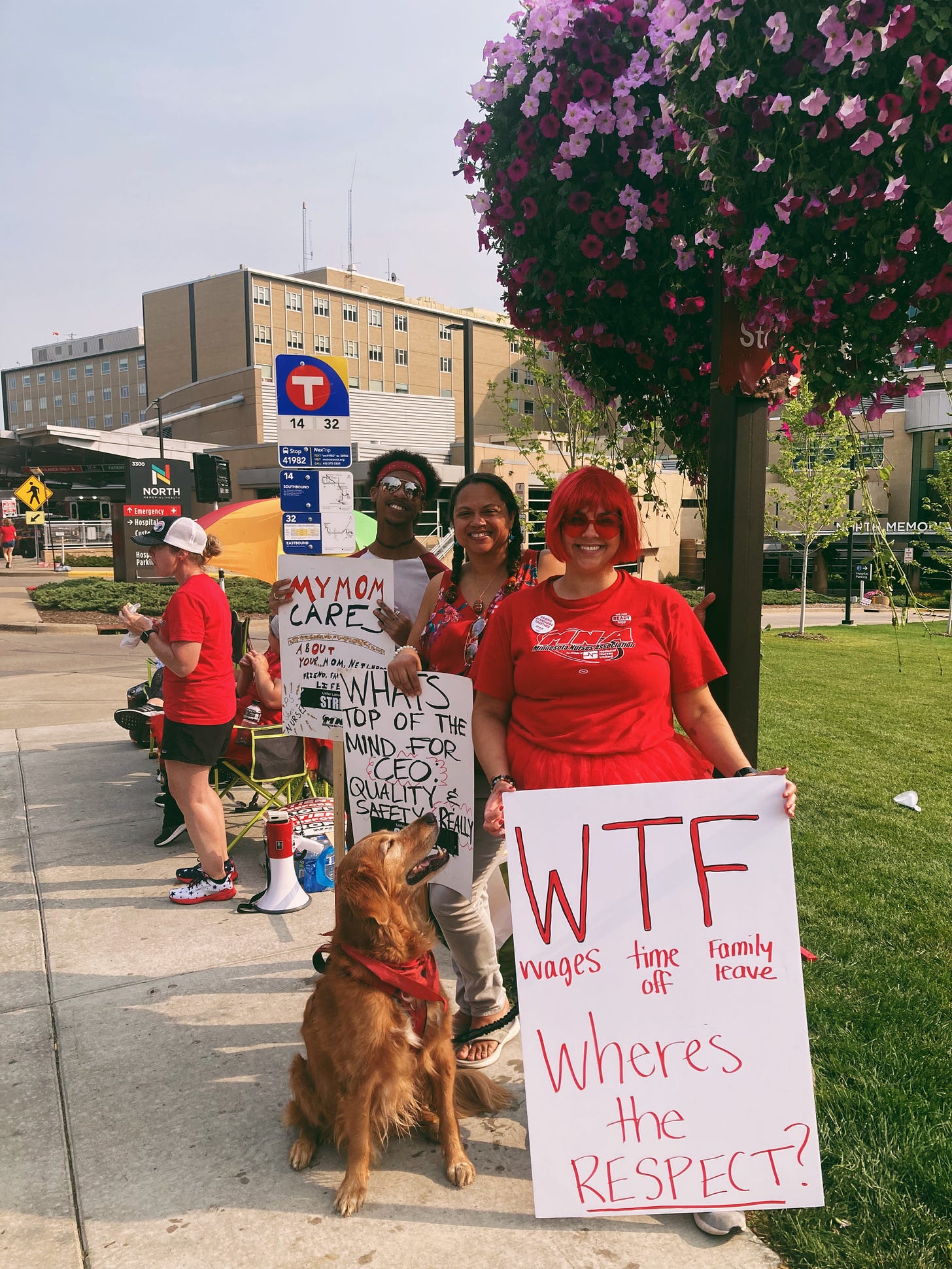 a few picketers dressed in red, one wearing a red wig, smile and hold signs. a golden retriever looks up at one of the picketers who holds a sign that reads "WTF: wages, time off, family leave"
