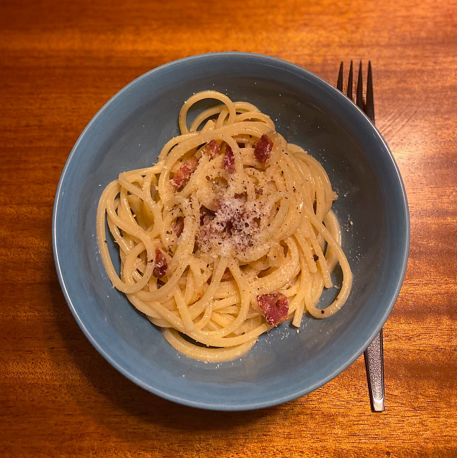 A blue bowl of spaghetti carbonara with grated pecorino and ground pepper on top.