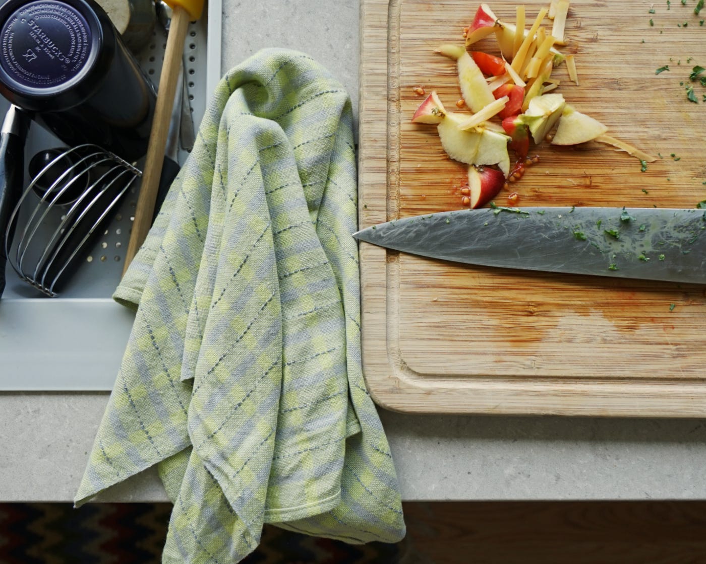 A folded cloth lying on a countertop. A cutting board and knife are to the right, with pieces of apple core, while a draining board with clean dishes is to the left