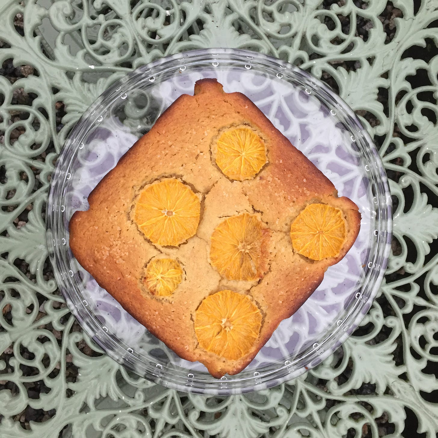 from above, a square cake with browned edges and slices of orange on top sits on a sea green wrought-iron patio table.